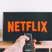 A person pointing a remote to a Netflix logo on a TV. Credit: Canva
