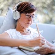 A woman listening to music on her headphones as she writes. Credit: Canva